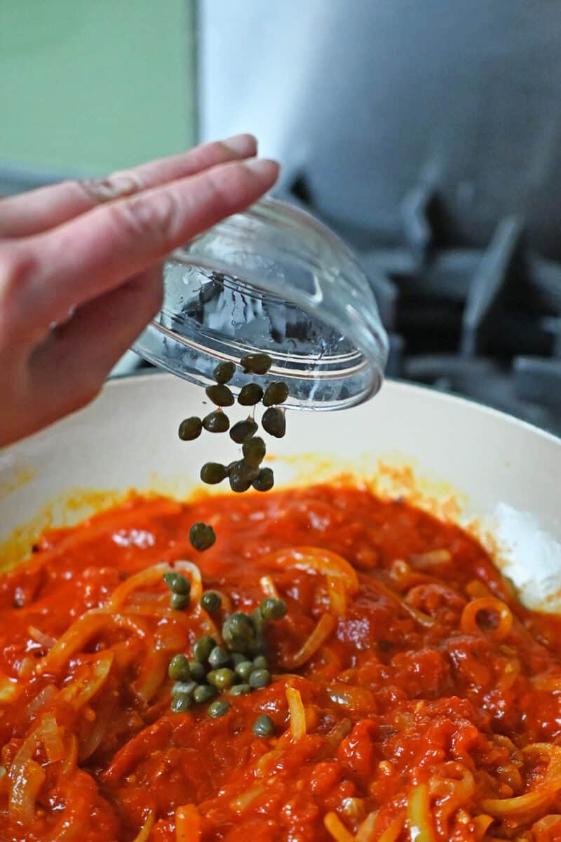A hand is adding some capers to a skillet filled with marinara sauce, sliced onions, minced garlic, and red pepper flakes.