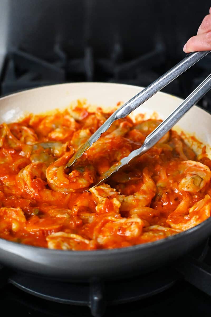 A pair of tongs are picking up fully cooked shrimp in an enamel cast iron skillet with spicy marinara sauce