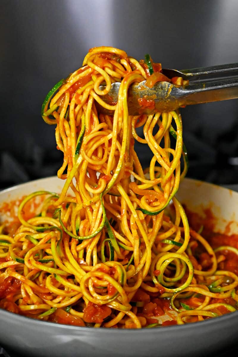 A pair of tongs tossing zucchini noodles in a skillet with spicy marinara sauce