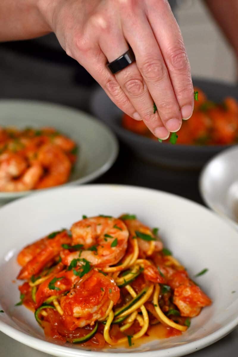 A hand is sprinkling minced Italian parsley on top of a bowl filled with paleo, Whole30, and keto spicy shrimp pasta