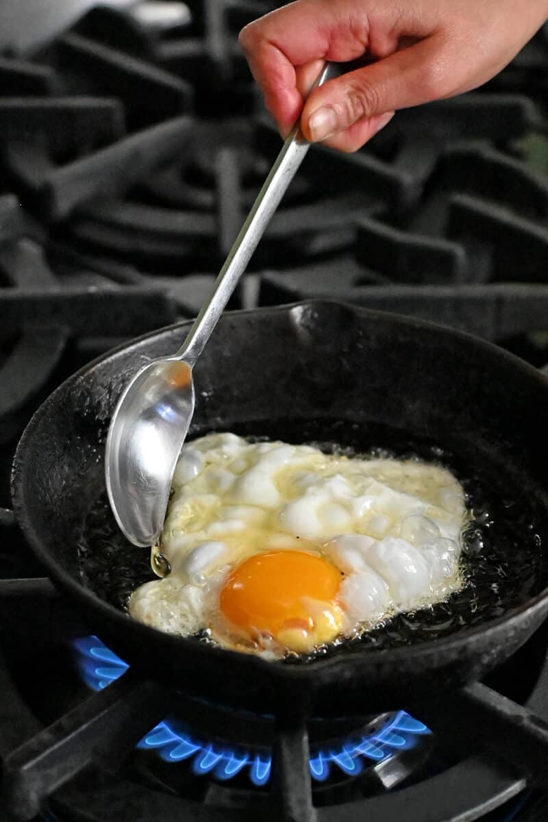 A spoon is basting a fried egg in a small cast iron skillet with hot oil.