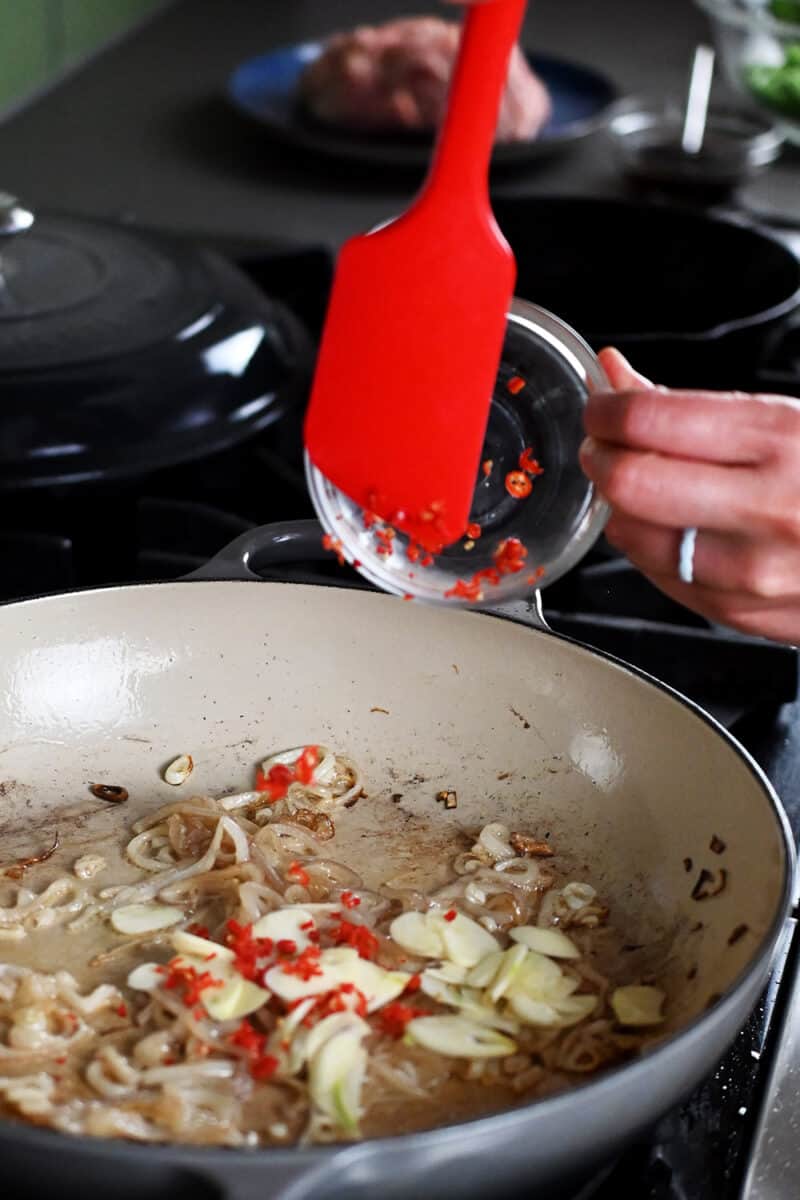 A red spatula is adding sliced garlic and thinly sliced Thai red peppers into a skillet with sautéed shallots.