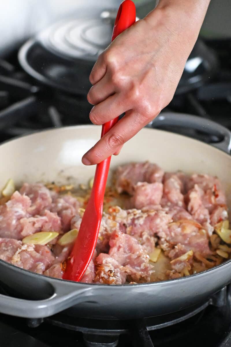 A red spatula is breaking up ground chicken into a skillet to make Thai Basil Chicken
