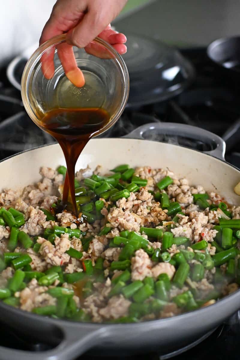 Pouring sauce into a skillet with ground chicken and green beans to make Thai Basil chicken