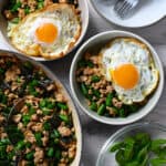 A skillet filled with Thai Basil Chicken is next to two bowls of it topped with fried eggs