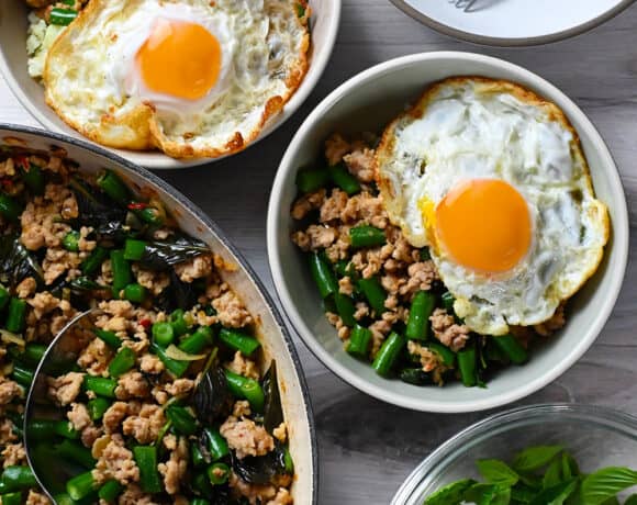 A skillet filled with Thai Basil Chicken is next to two bowls of it topped with fried eggs