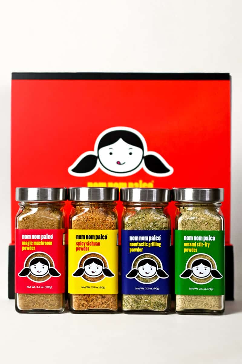 Photograph of the Nom Nom Paleo spice blend collection: four spice bottles in front of a red collector's box with the Nom Nom Paleo logo