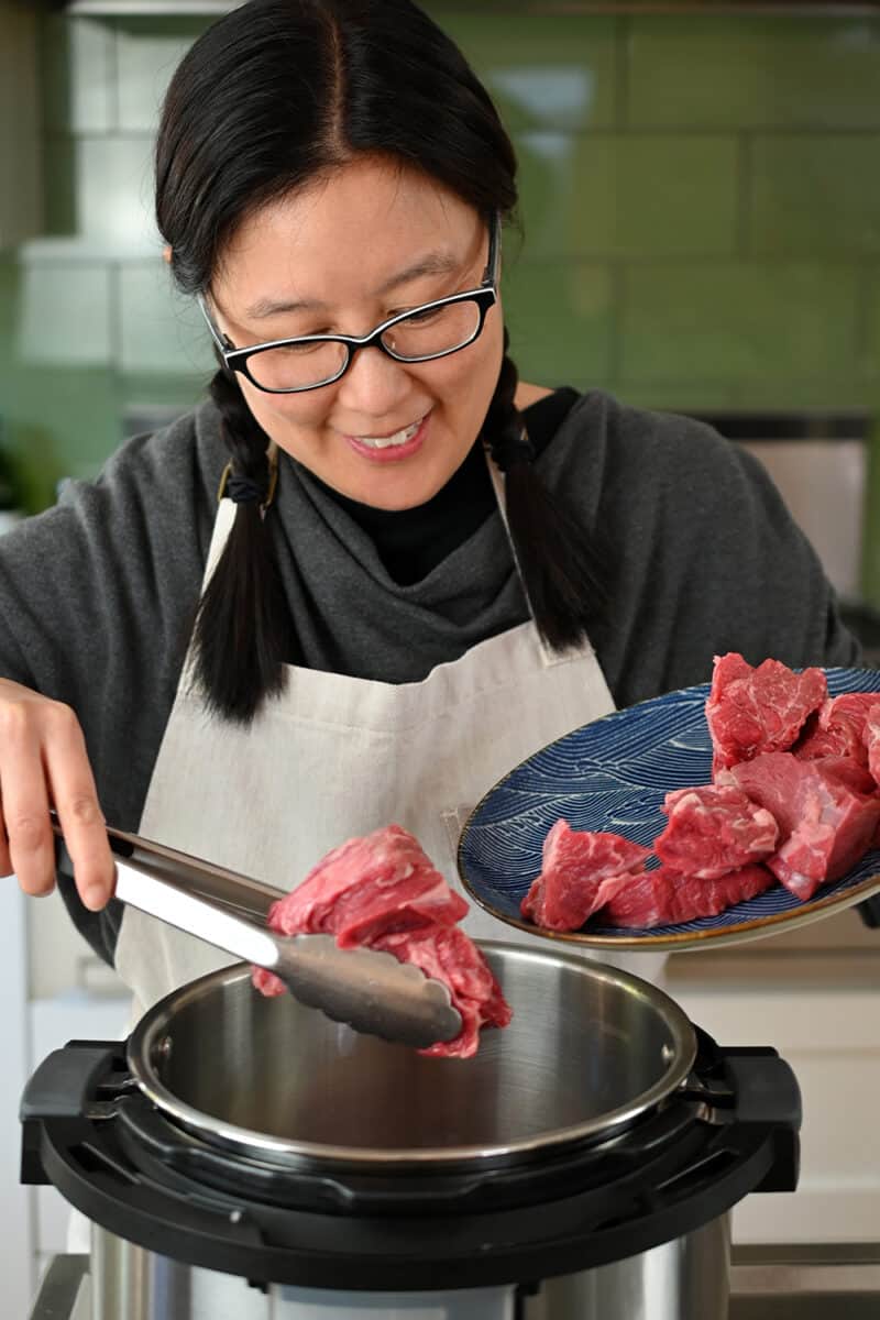 An Asian woman in glasses is adding beef cubes to an open Instant Pot