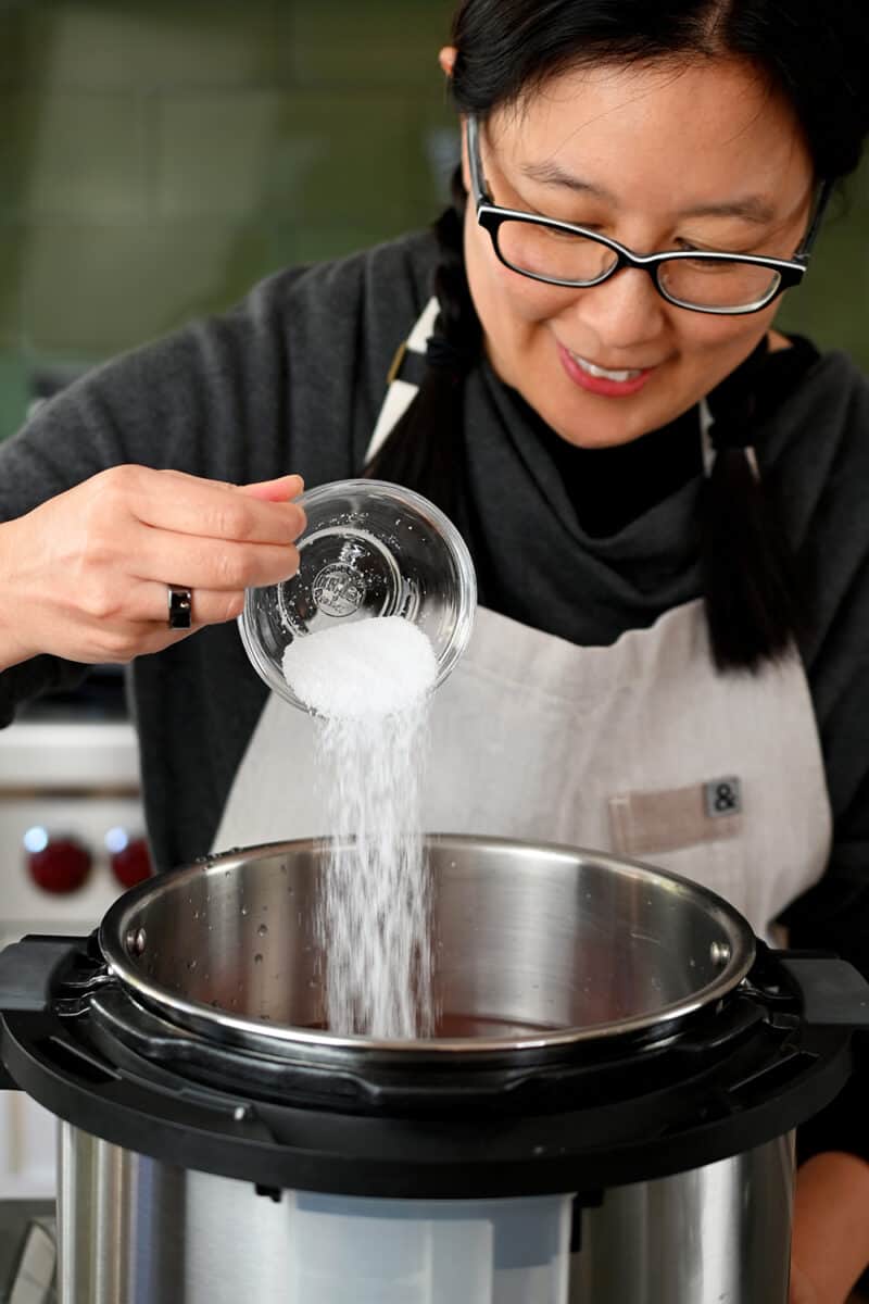 An Asian woman in glasses is adding some salt to an open Instant Pot to make Chinese Braised Beef Noodle Soup