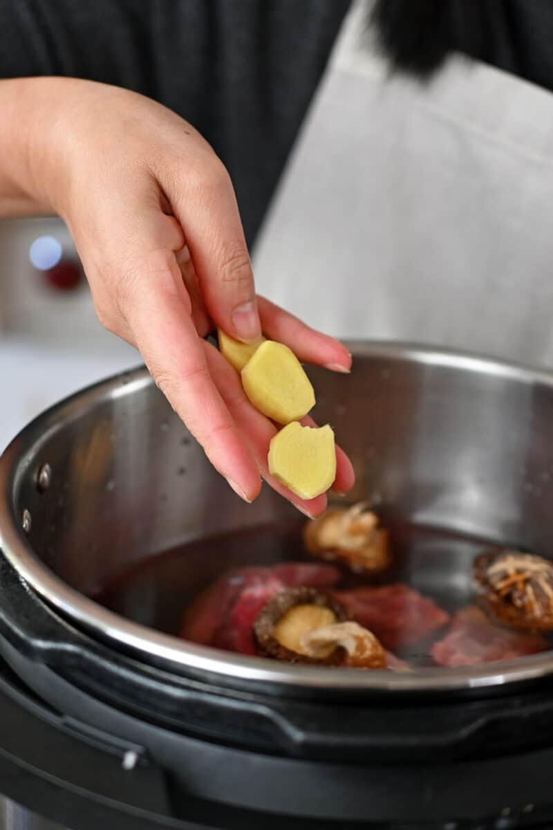 Adding sliced ginger to an open Instant Pot filled with ingredients to make Chinese Beef Noodle Soup