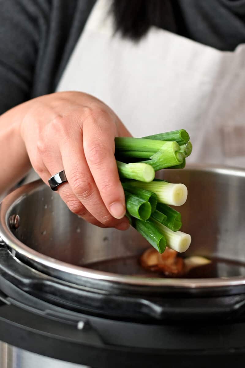 A hand is adding scallions to an open Instant Pot filled with Chinese Beef Noodle Soup ingedients
