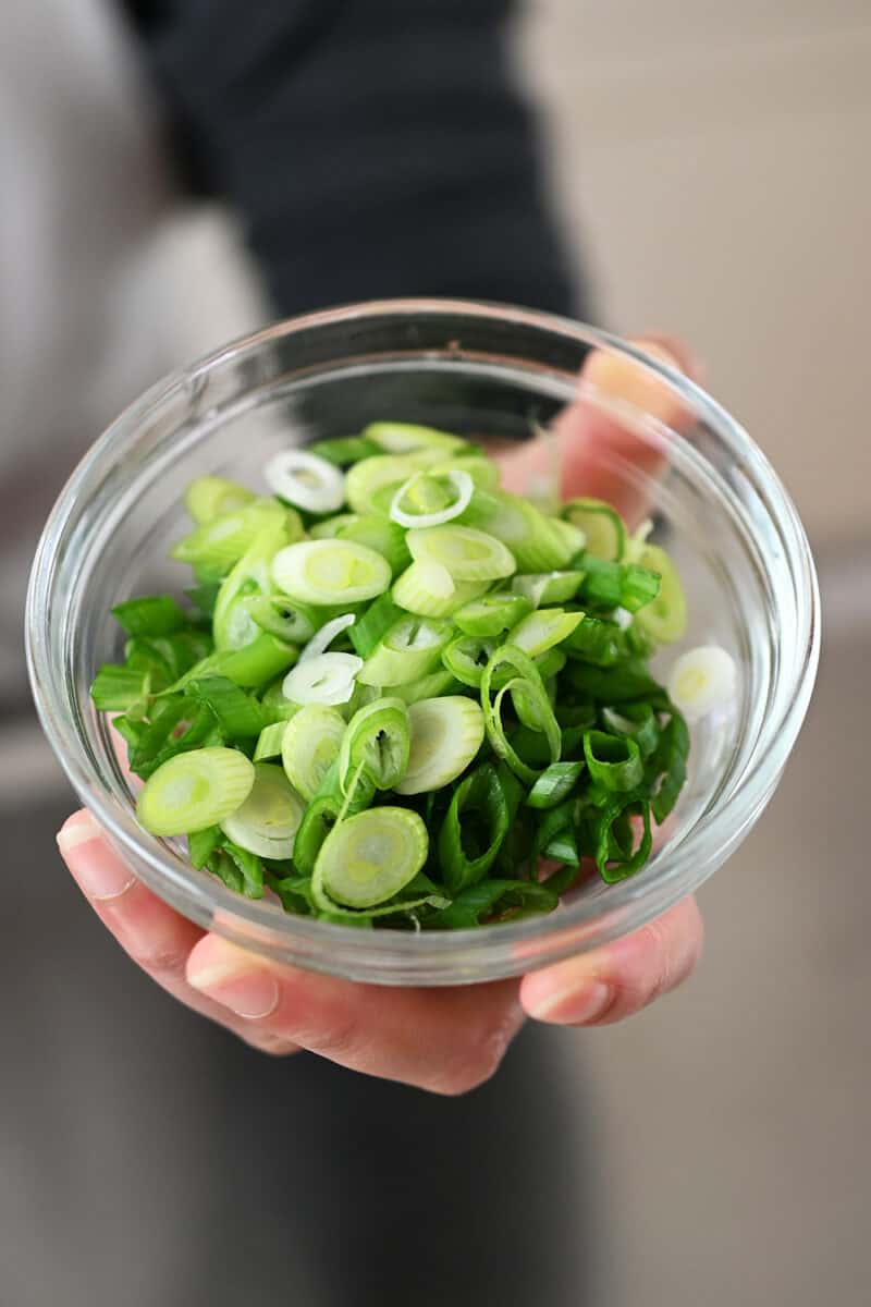 A hand showing a small glass bowl filled with thinly sliced scallions