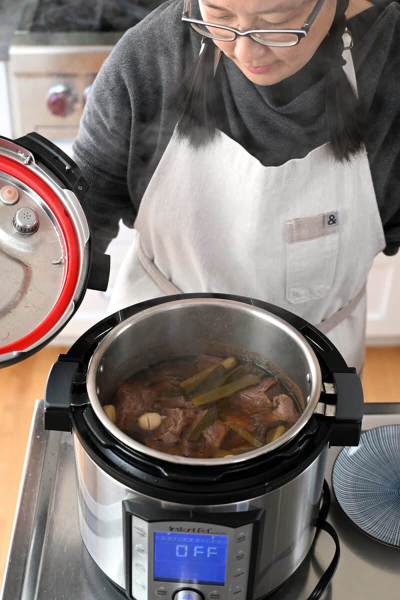 An Asian woman taking the lid off an Instant Pot filled with Chinese Beef Noodle soup