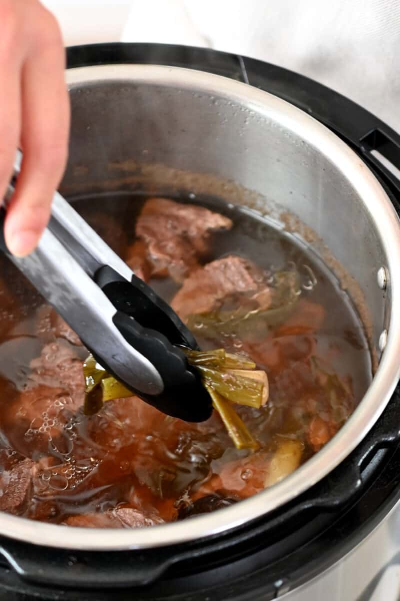 A pair of tongs is removing sliced ginger from an open Instant Pot filled with beef broth