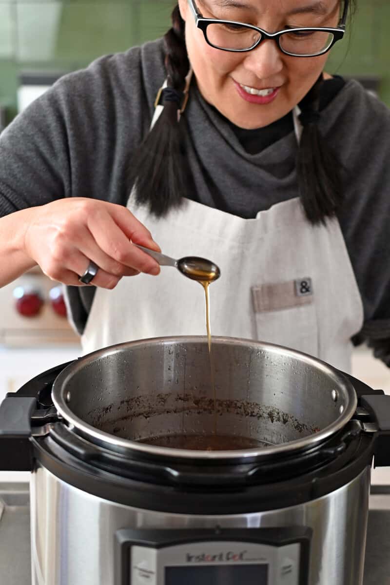 A smiling Asian woman is adding a spoonful of sesame oil to an Instant Pot filled with beef broth.