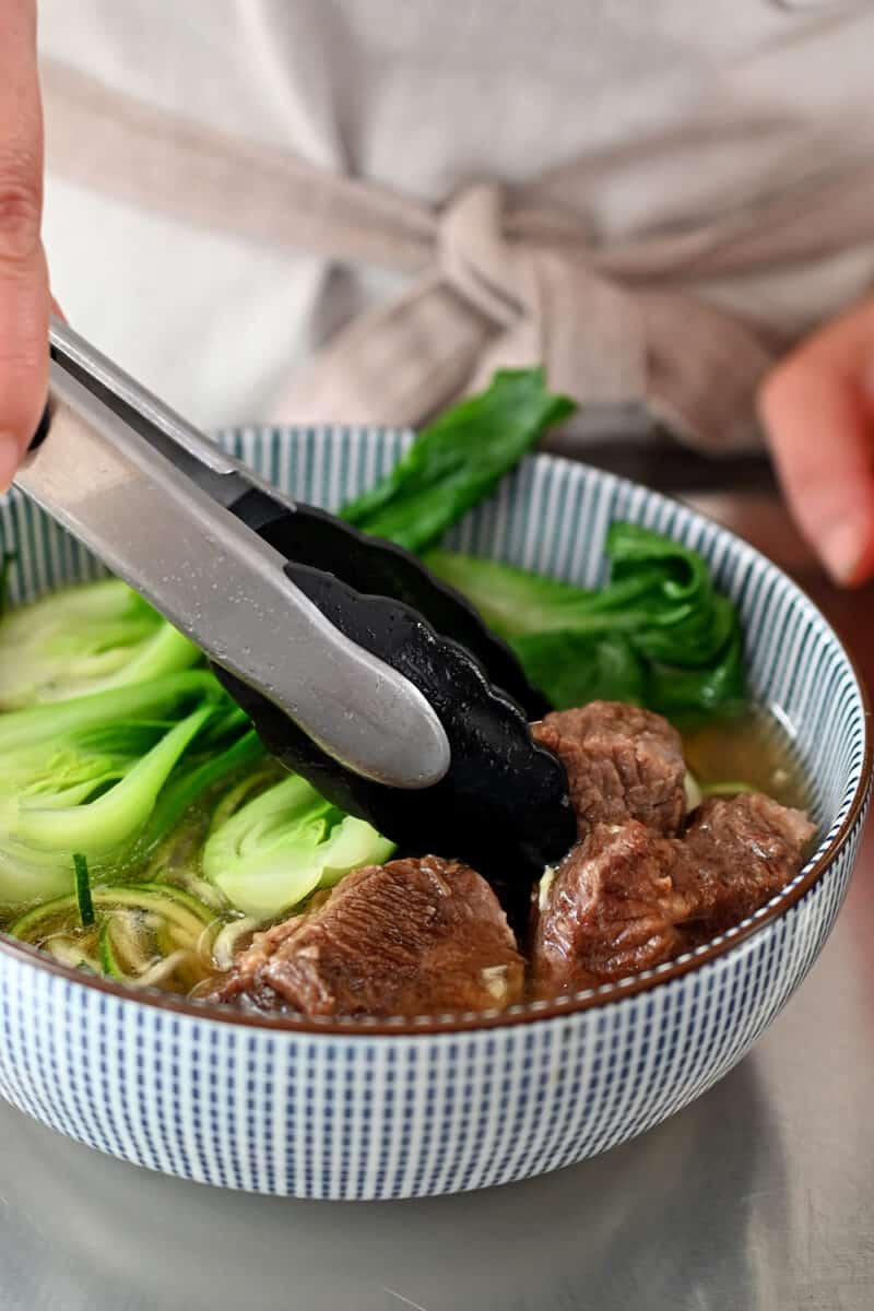 A pair of tongs is adding stewed beef cubes on a bowl of Chinese beef noodle soup
