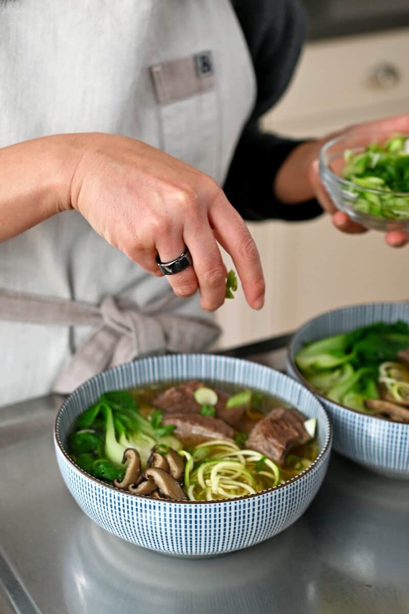 A hand is sprinkling sliced scallions on a bowl of paleo, Whole30, gluten-free, and keto Chinese Beef Noodle Soup