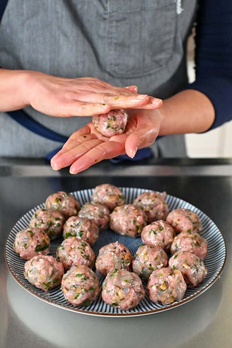 Someone in a gray apron is using their hands to roll wonton meatballs and placing them on a blue and white plate.