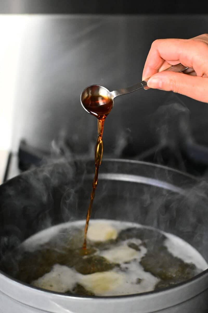 Pouring a spoonful of fish sauce into a simmering pot of chicken broth and ginger.