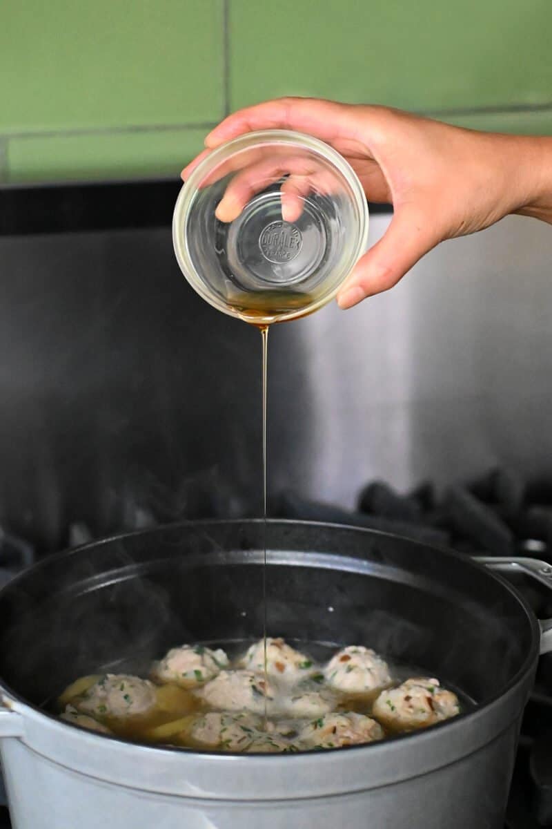 A hand is adding toasted sesame oil from a small bowl to a pot of wonton meatball soup