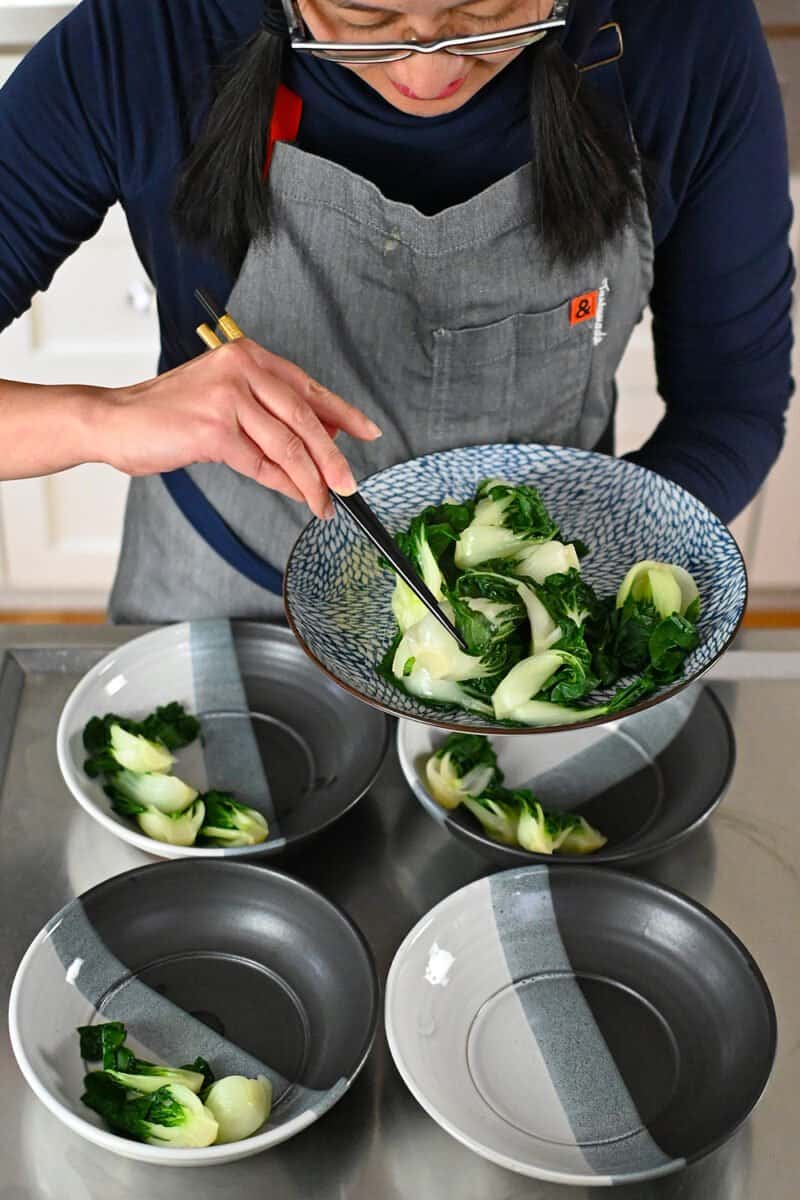 A woman is dividing cooked baby Bok Choy into four soup bowls.