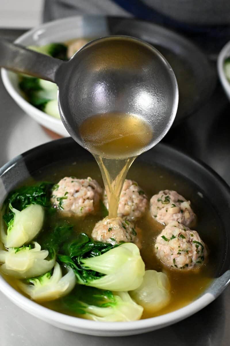 A ladle is adding broth to a bowl filled with wonton meatballs and baby Bok Choy.