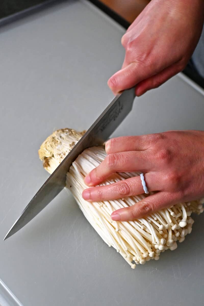 A chef's knife is cutting the root ends off of a bunch of enoki mushrooms