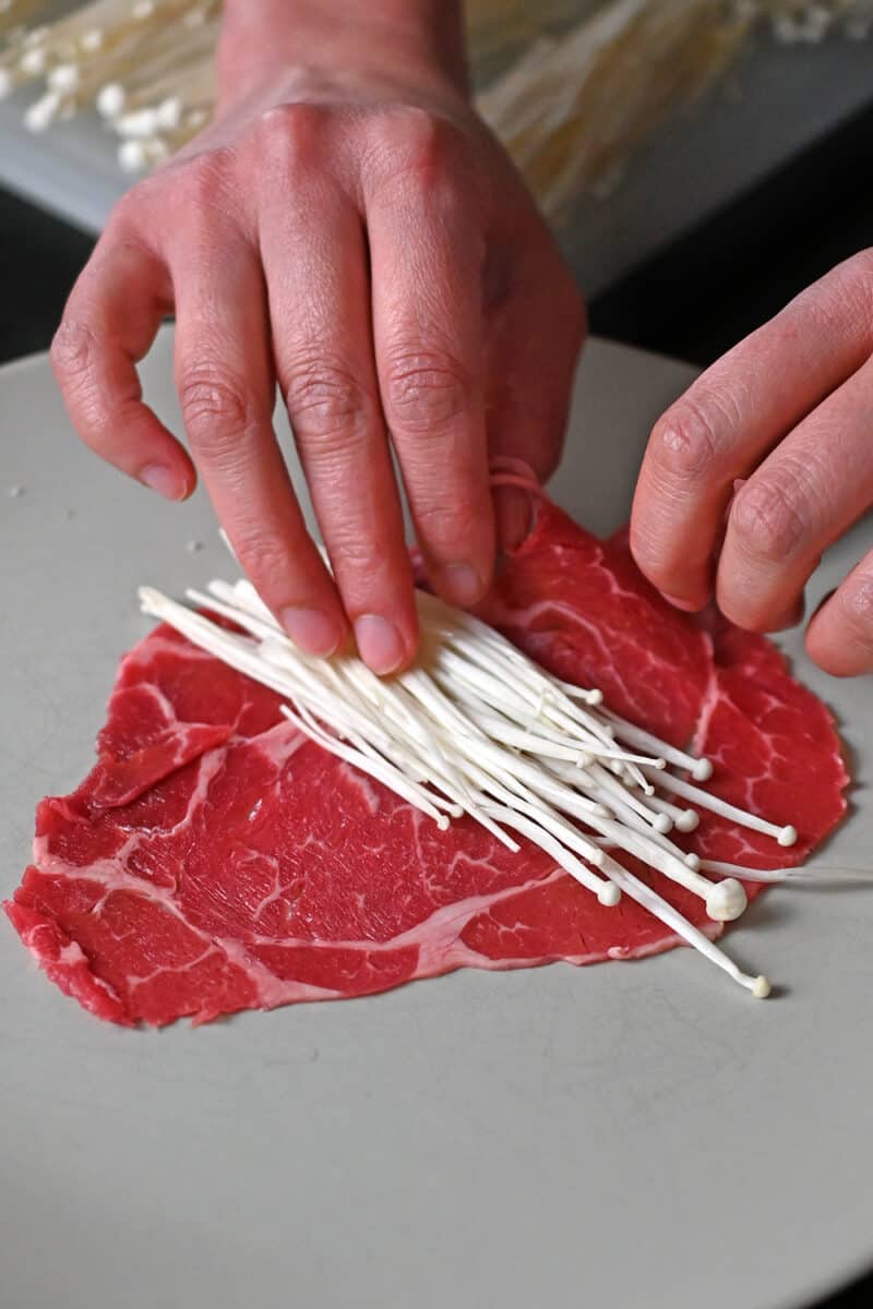 A small stack of enoki mushrooms is on a paper thin slice of beef