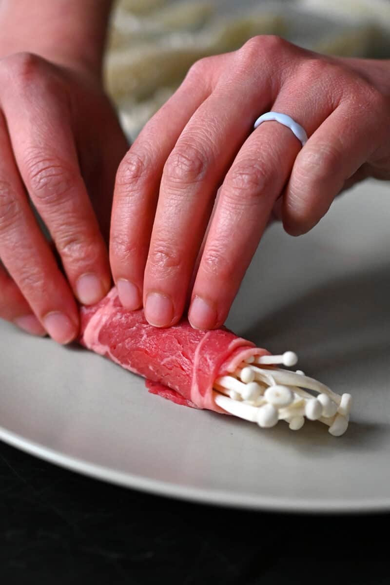 Two hands are rolling enoki mushrooms in a thin slice of raw beef.