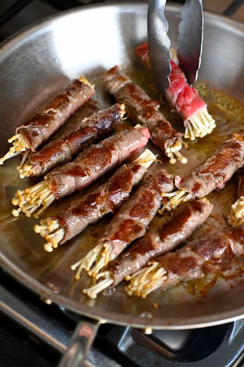 Golden brown beef and enoki rolls frying in a large skillet 