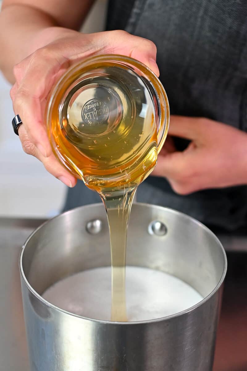 Adding honey from a small bowl into a small saucepan filled with coconut milk and coconut water.