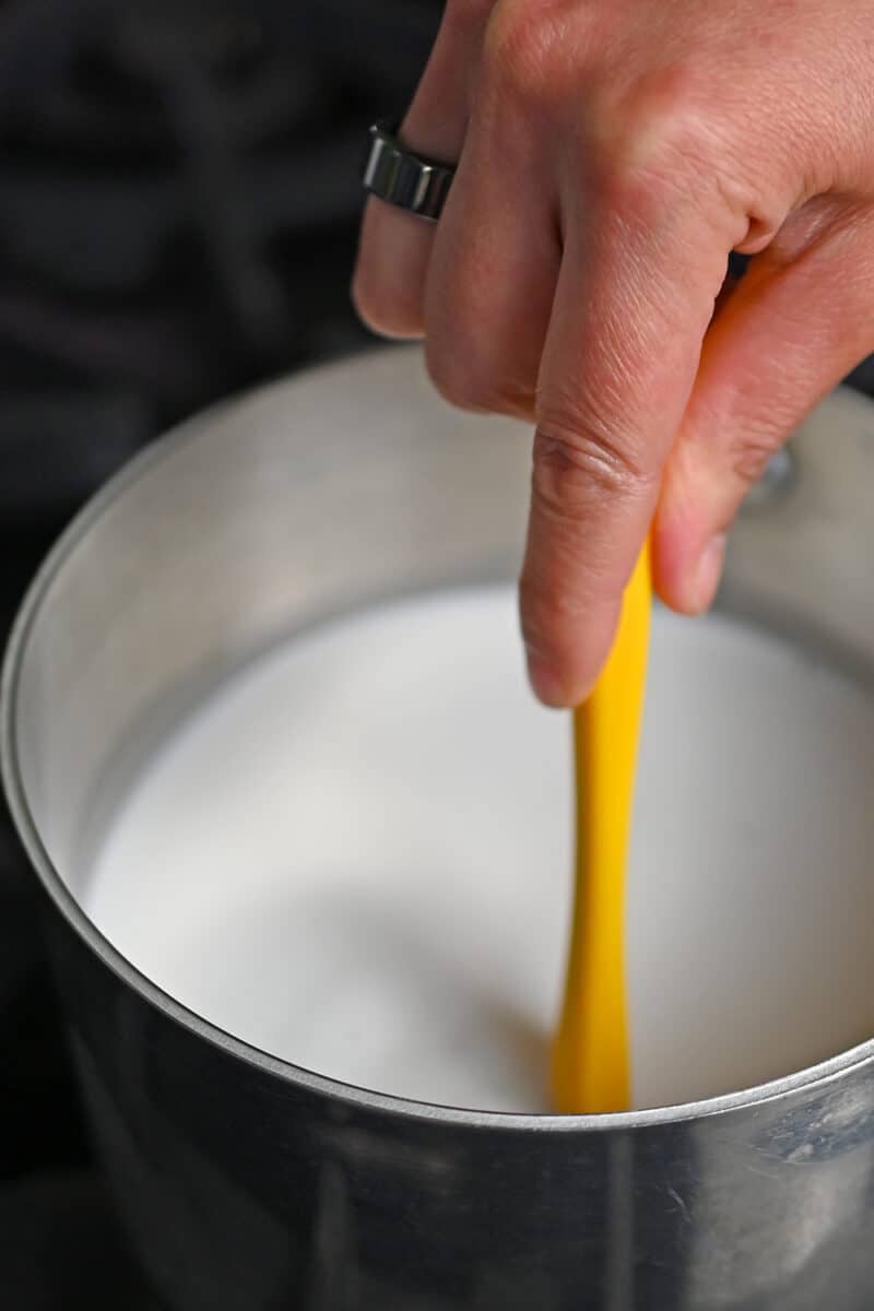 A hand is mixing the coconut mixture in a saucepan with a yellow spatula.