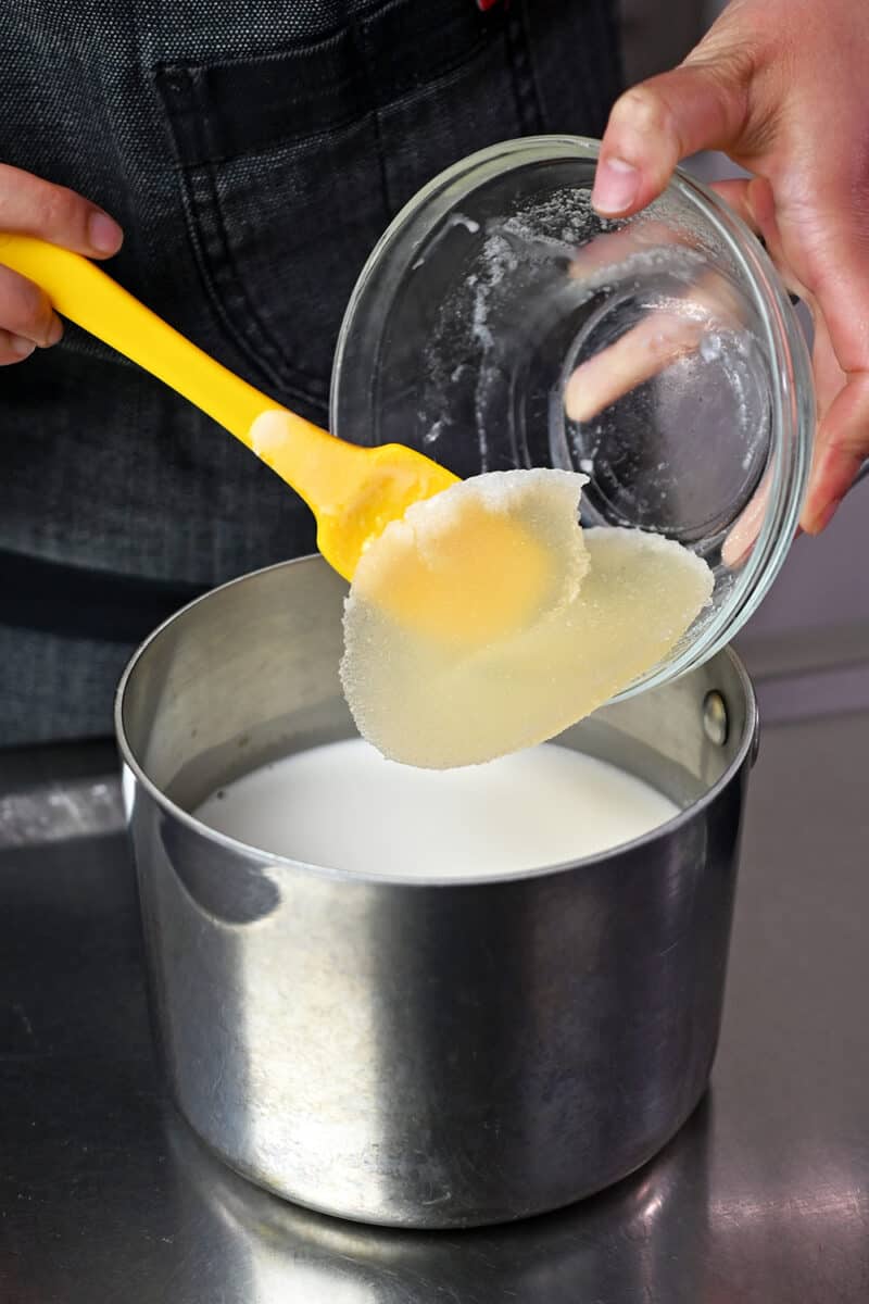 A yellow spatula is adding rehydrated gelatin into a saucepan filled with coconut milk and coconut water.