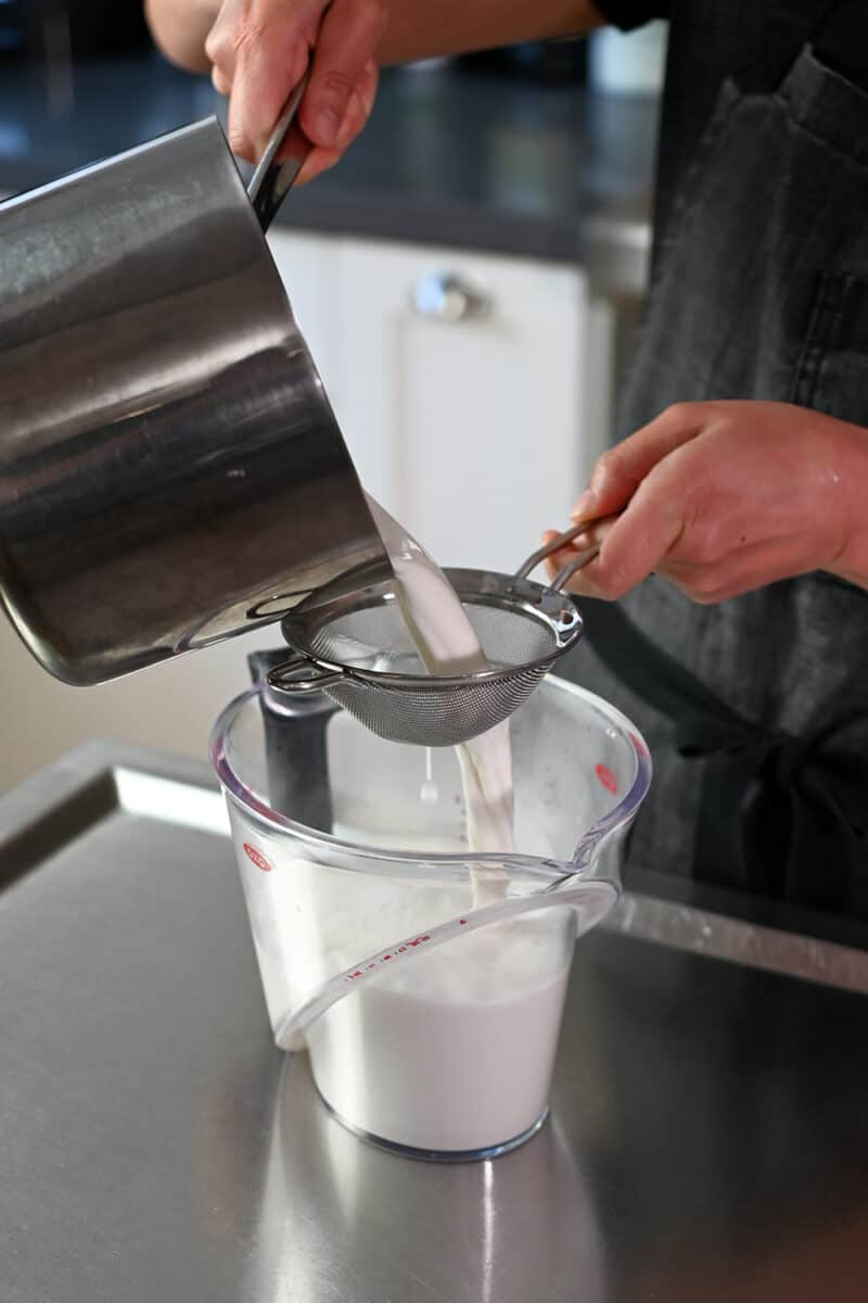 The hot coconut milk mixture is poured from the saucepan into a liquid measuring cup through a fine mesh sieve.
