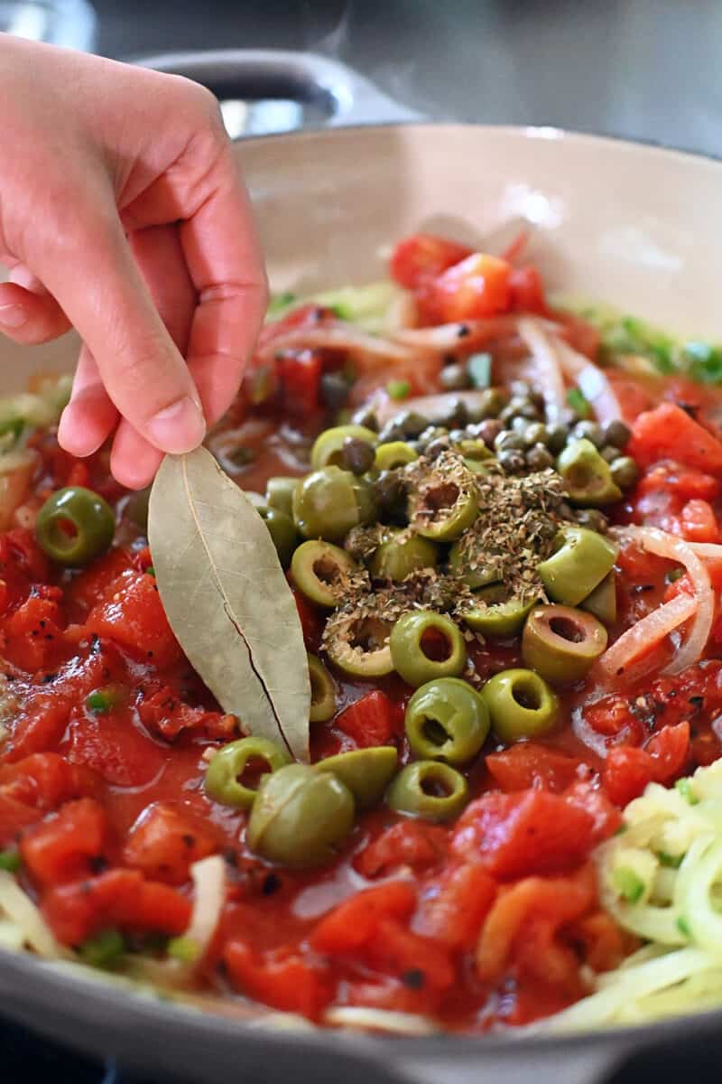 A hand is adding a dried bay leaf to a skillet with canned diced tomatoes, sliced green olives, capers, and dried oregano to make homemade Veracruz sauce