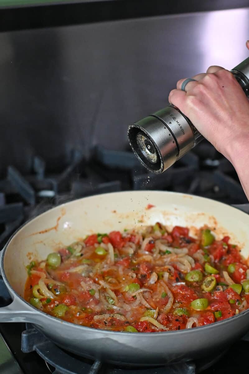 A hand is adding freshly cracked pepper to a skillet filled with homemade Veracruz sauce