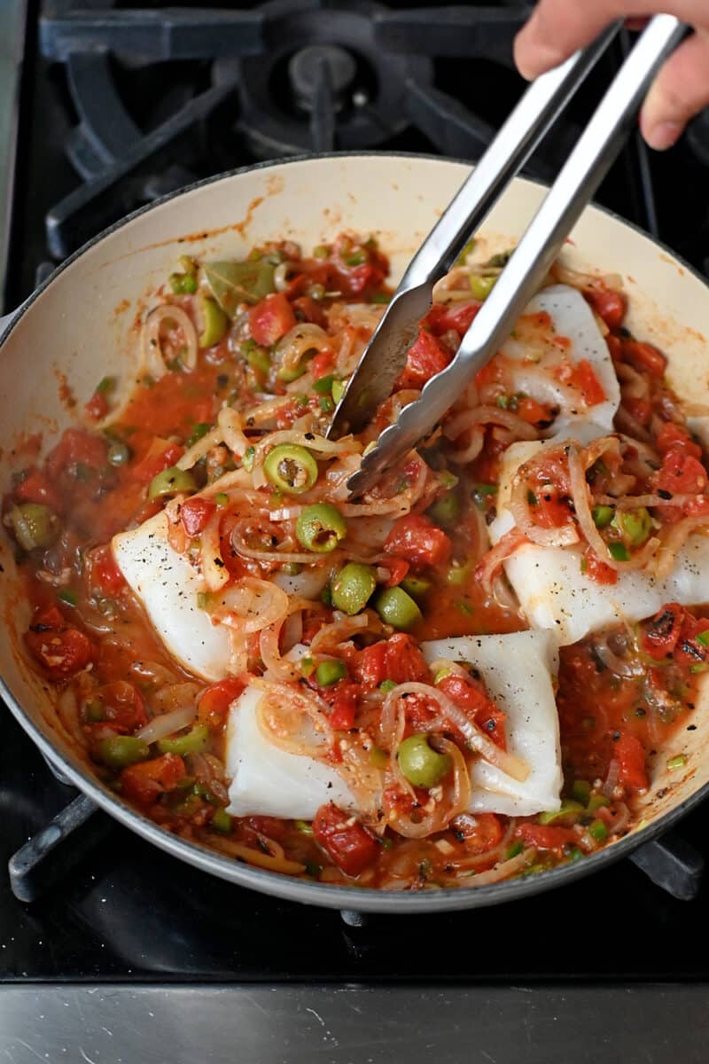 A pair of tongs is tucking raw fish fillets into a skillet filled with Veracruz sauce.