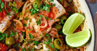 An overhead shot of white fish Veracruz in a skillet with sliced lime wedges on the side.