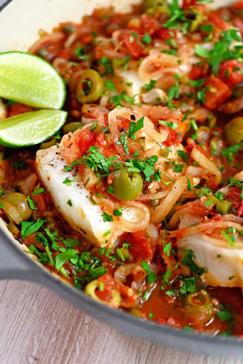 A closeup of white fish filets in Veracruz sauce with lime wedges on the side.