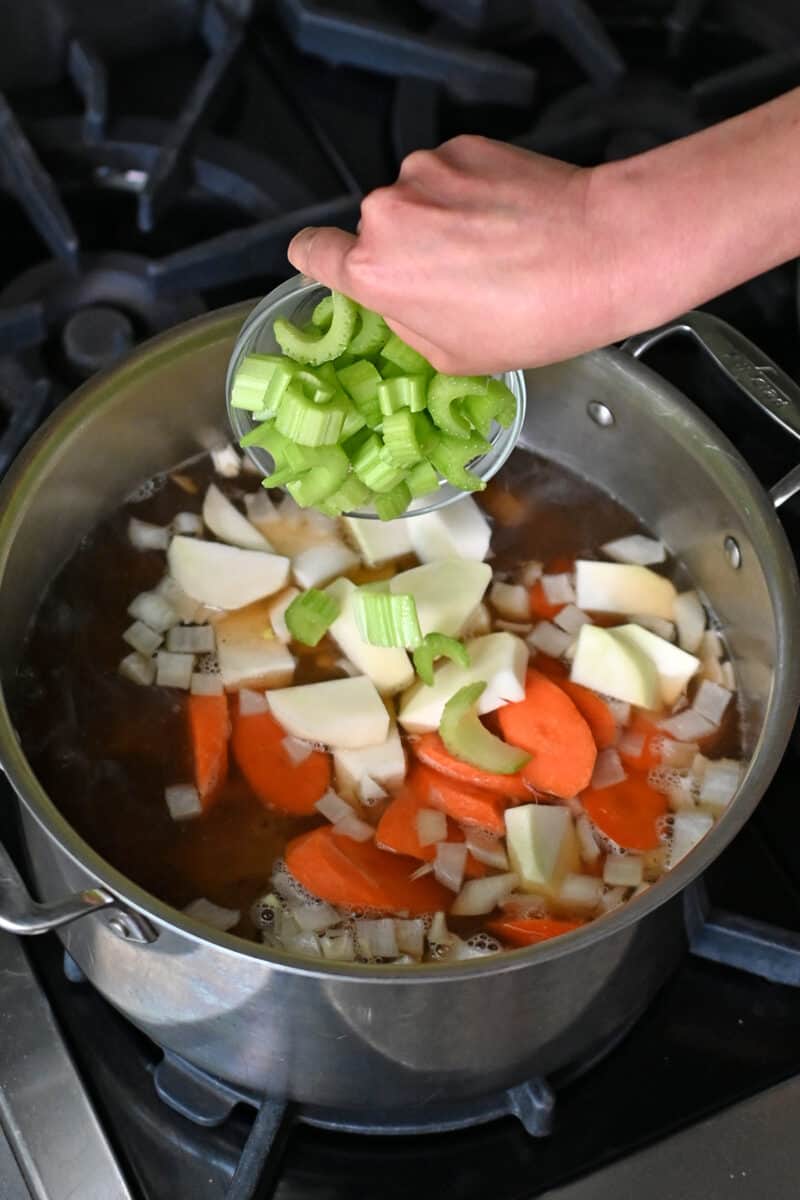 Adding carrots, onions, kohlrabi, and celery to a pot filled with oxtail soup