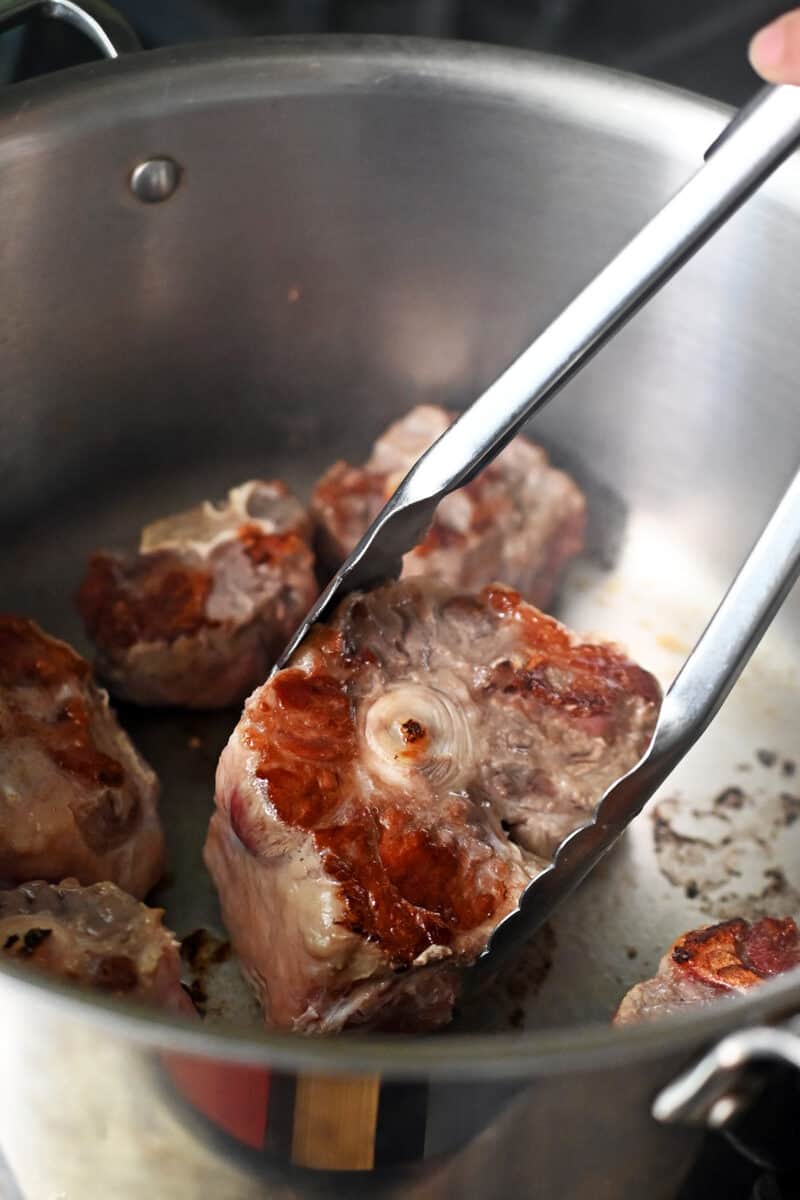 A pair of tongs is flipping a seared oxtail in a large pot.