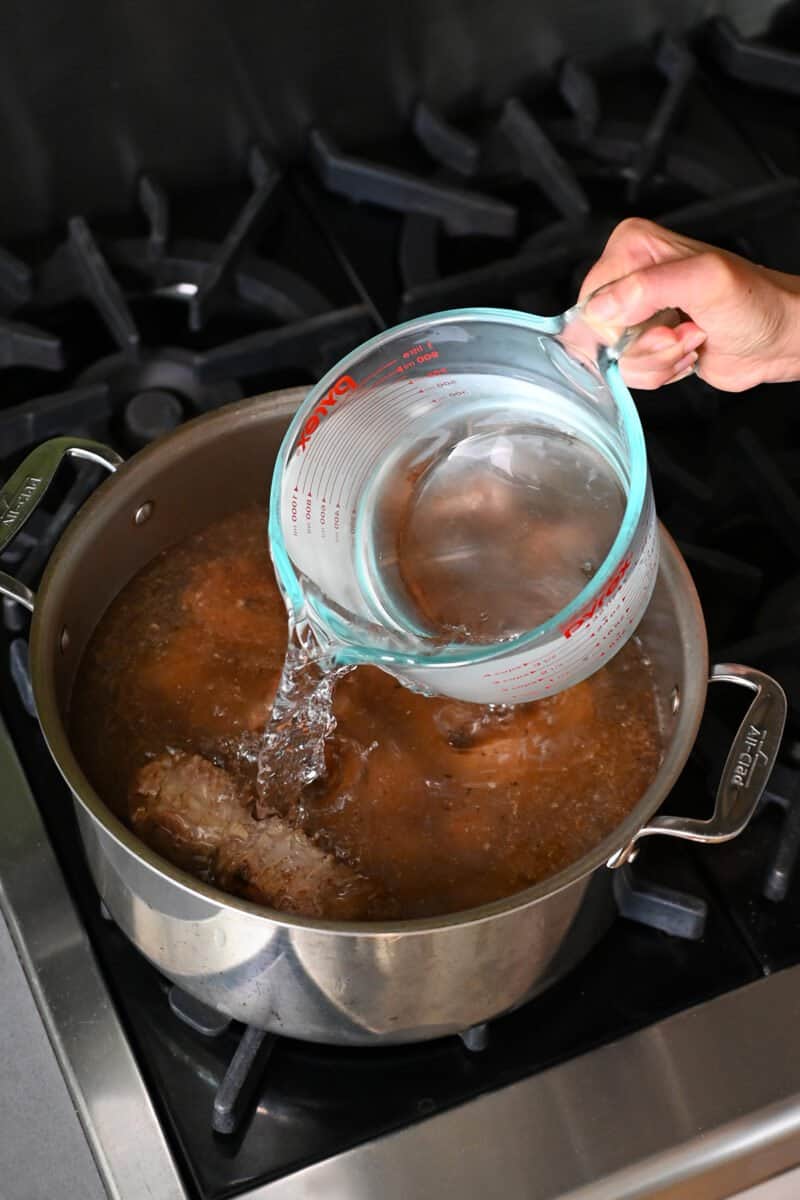 Pouring water into a large pot filled with seared oxtails.