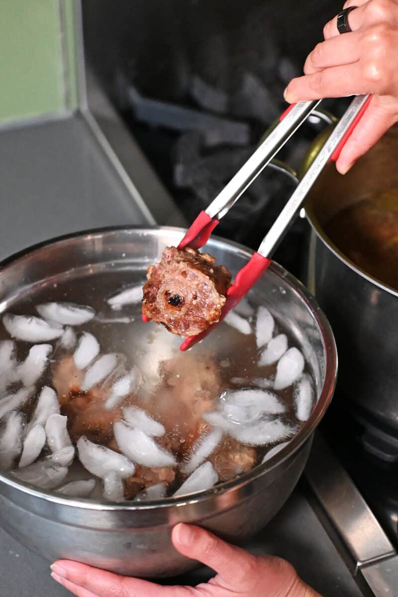 A pair of tongs is transferring the cooked oxtail into an ice water bath.