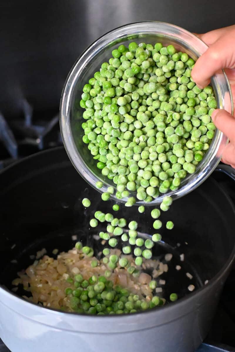 Pouring a medium bowl filled with frozen green peas into a pot filled with sautéed chopped shallots and garlic.