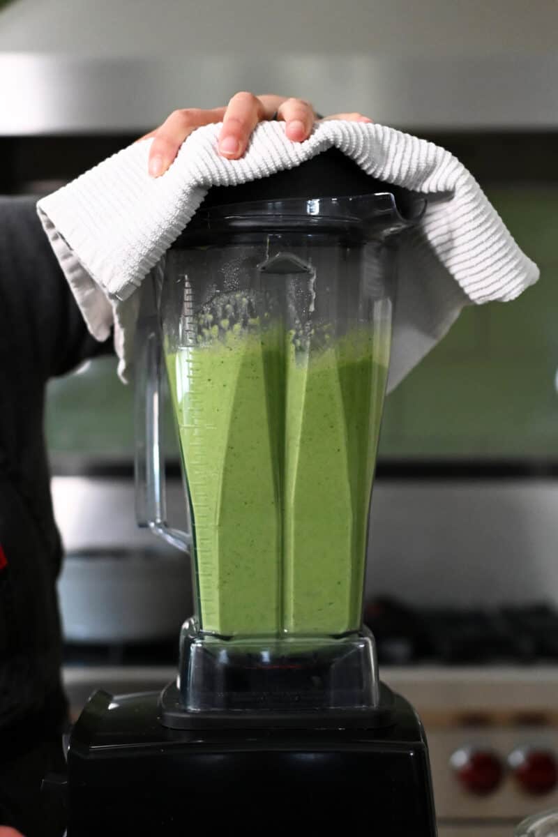 A side view of a Vitamix blender filled with hot green pea soup. The lid has a towel on top.