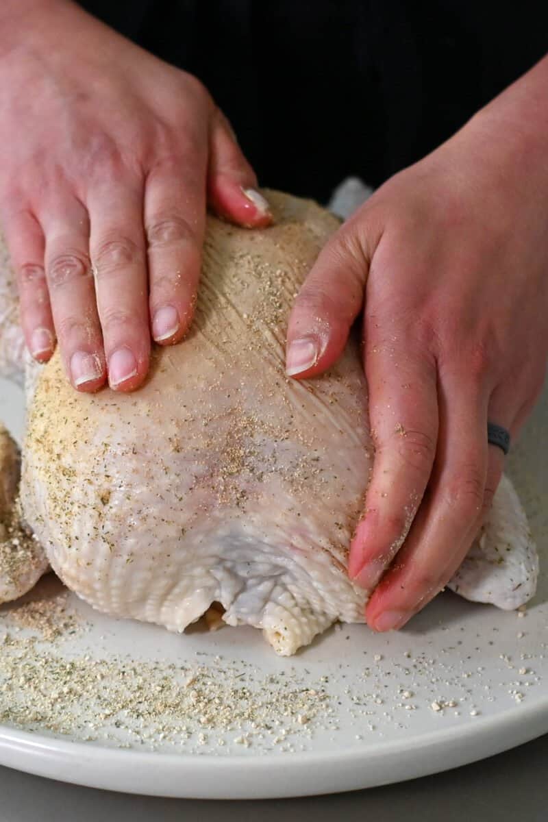 Two hands are rubbing a seasoning blend on top of a raw whole chicken.