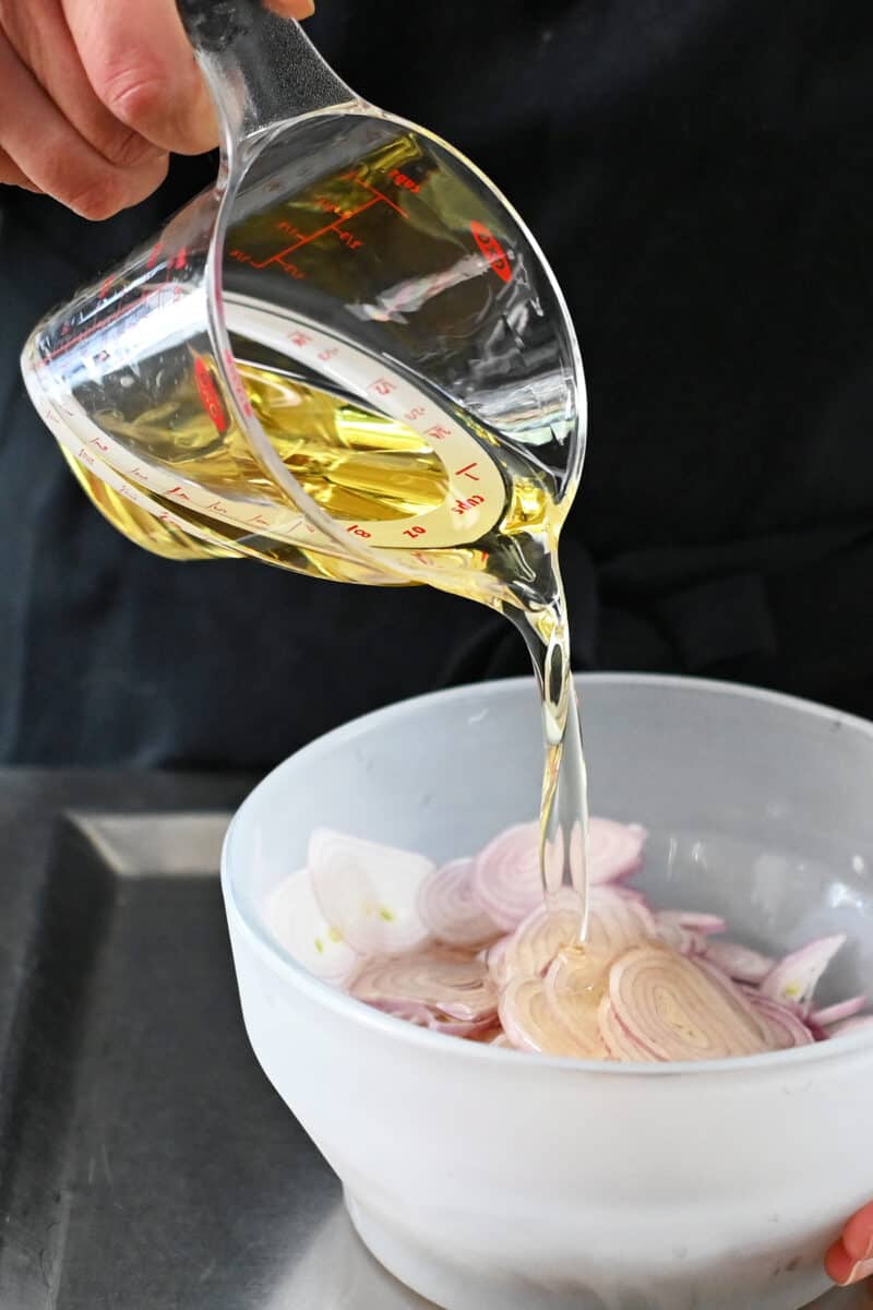 Pouring avocado oil from a liquid measuring cup into a glass bowl filled with sliced shallots.