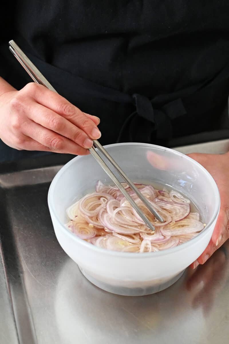 Someone using stainless steel chopsticks to submerge sliced shallots in oil.