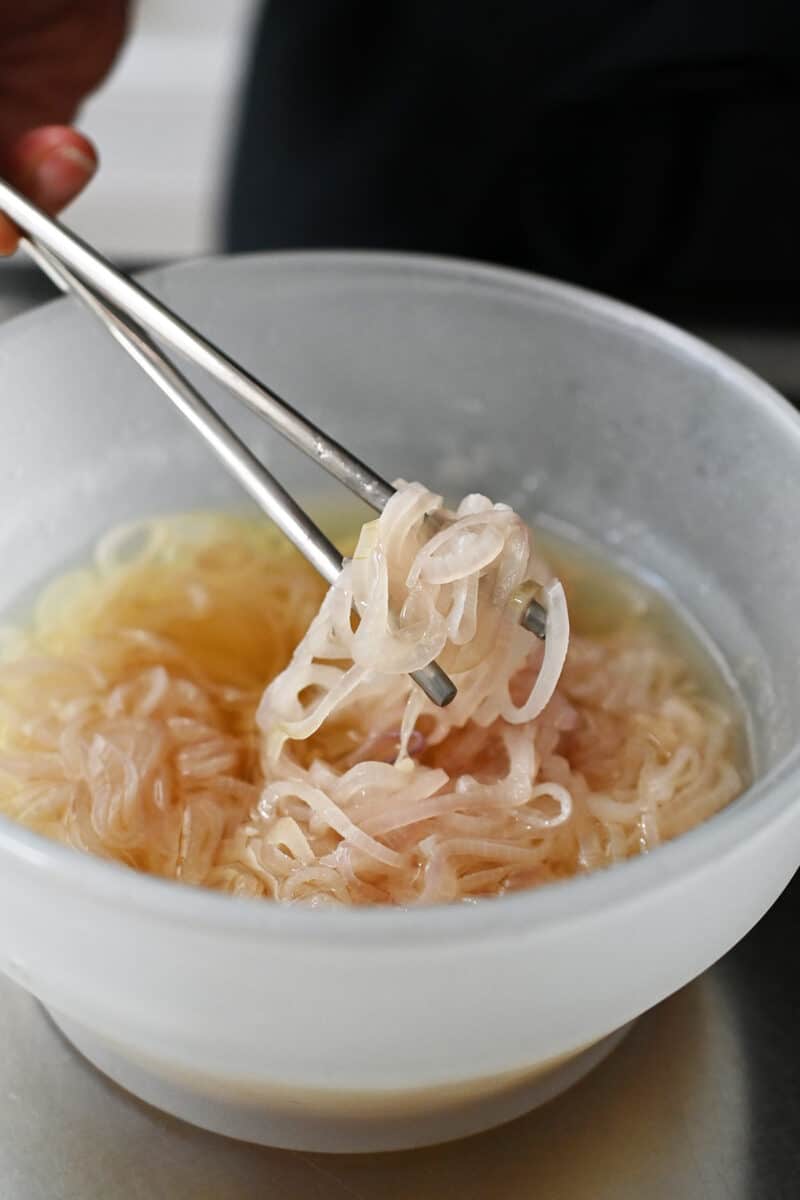 A pair of chopsticks is lifting some softened sliced shallots out of a bowl filled with avocado oil.