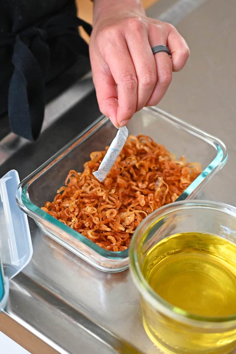 A hand is adding a desiccant packet to a glass container with crispy shallots.